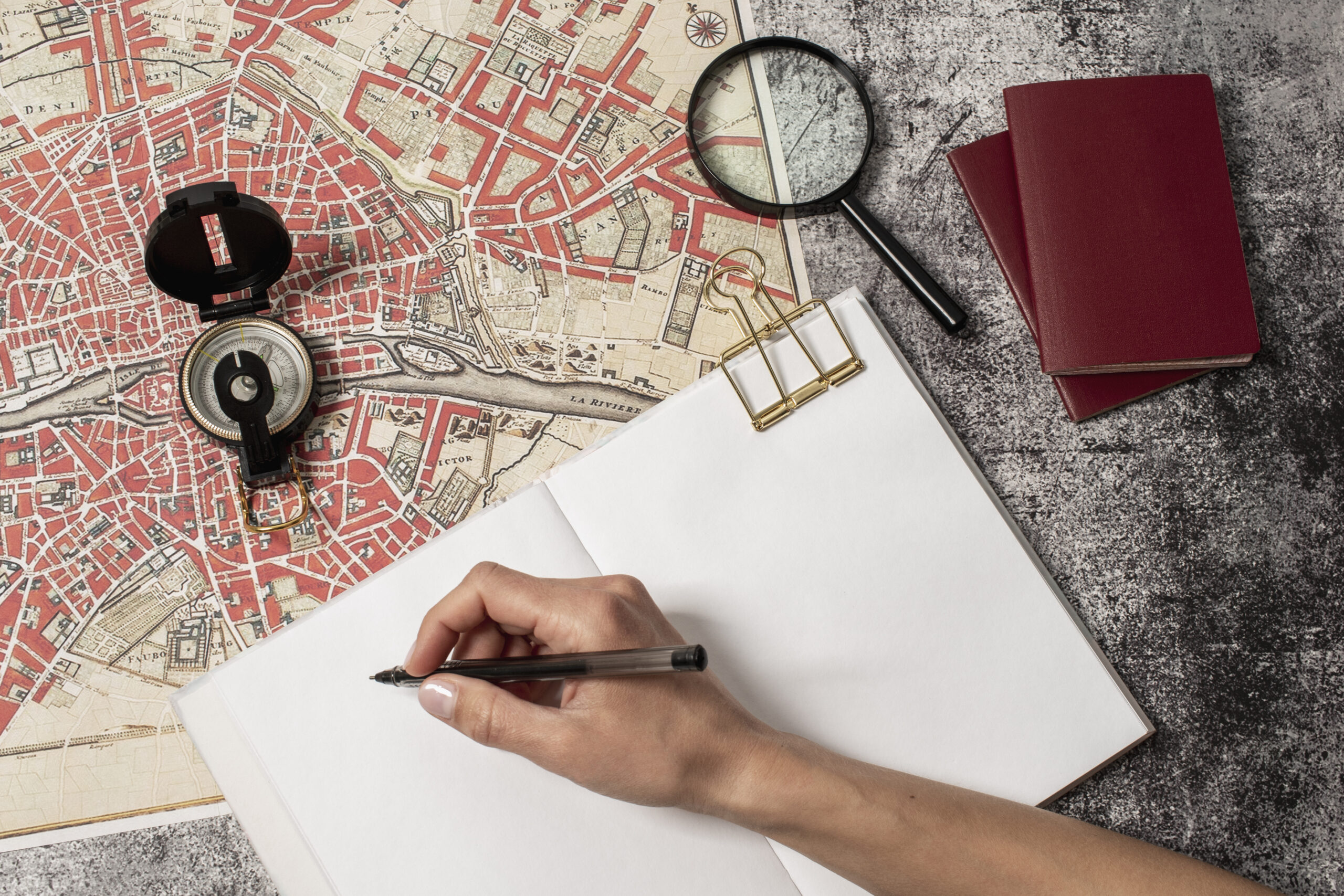 Na imagem, aparece uma mão com uma caneta escrevendo em um papel ao lado de um mapa e lupa